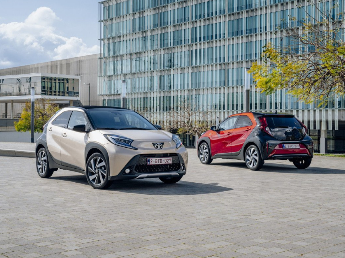 Dos Toyota Aygo X Cross aparcados en una plaza urbana, uno de color crema visto de frente y otro rojo visto desde la parte trasera, con edificios modernos al fondo.