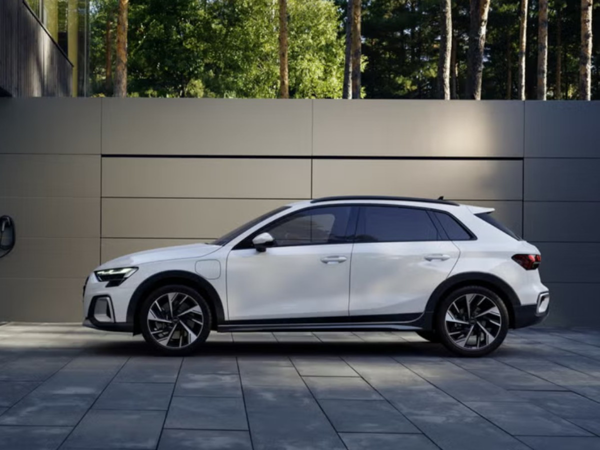 Audi A3 Allstreet blanco visto de perfil, aparcado en una zona exterior moderna con suelo de losas grises y un muro liso, rodeado de árboles al fondo.