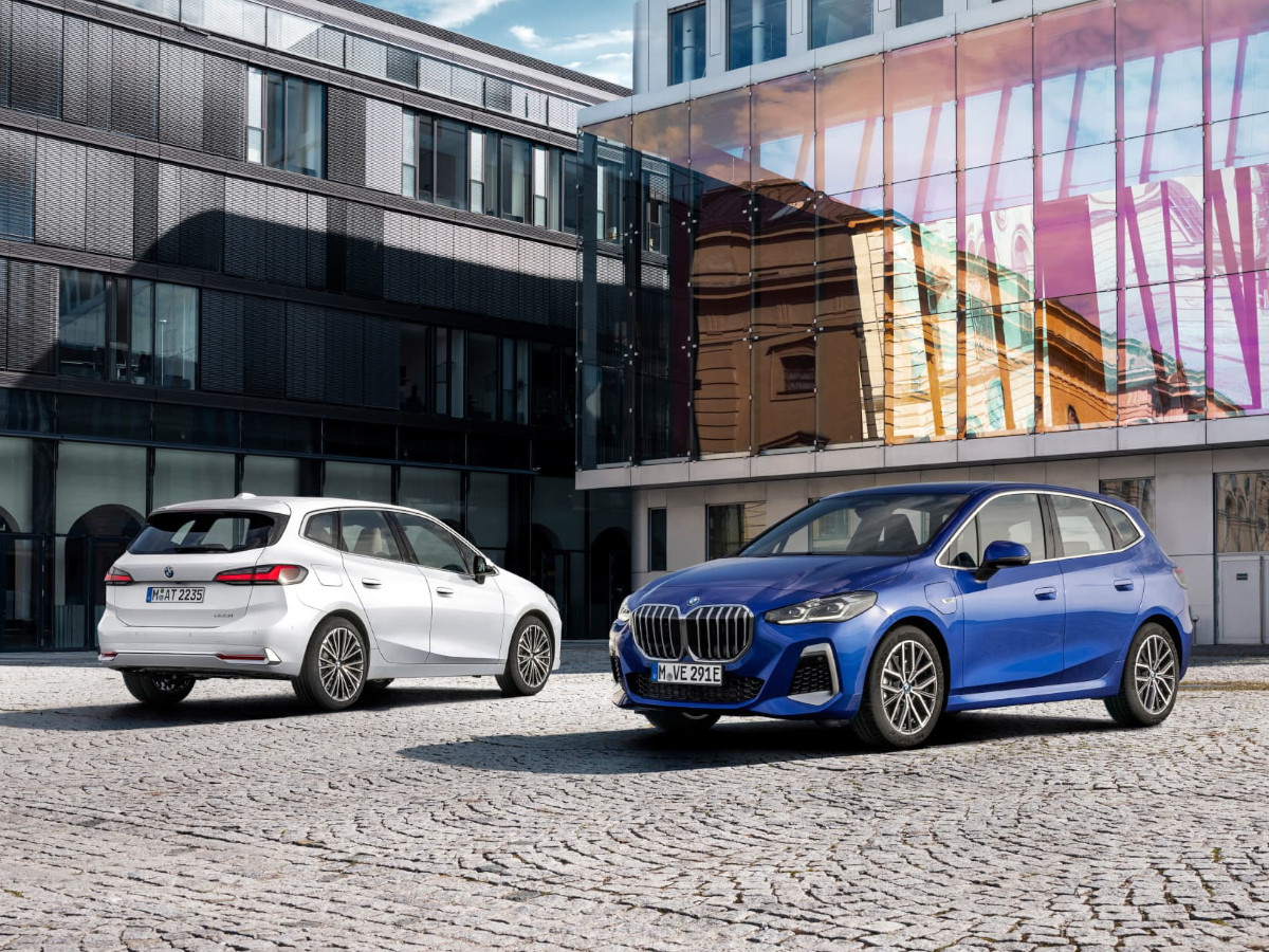 Dos BMW Serie 2 Active Tourer, uno blanco y otro azul, estacionados en una plaza urbana con edificios modernos de cristal y ladrillo al fondo.