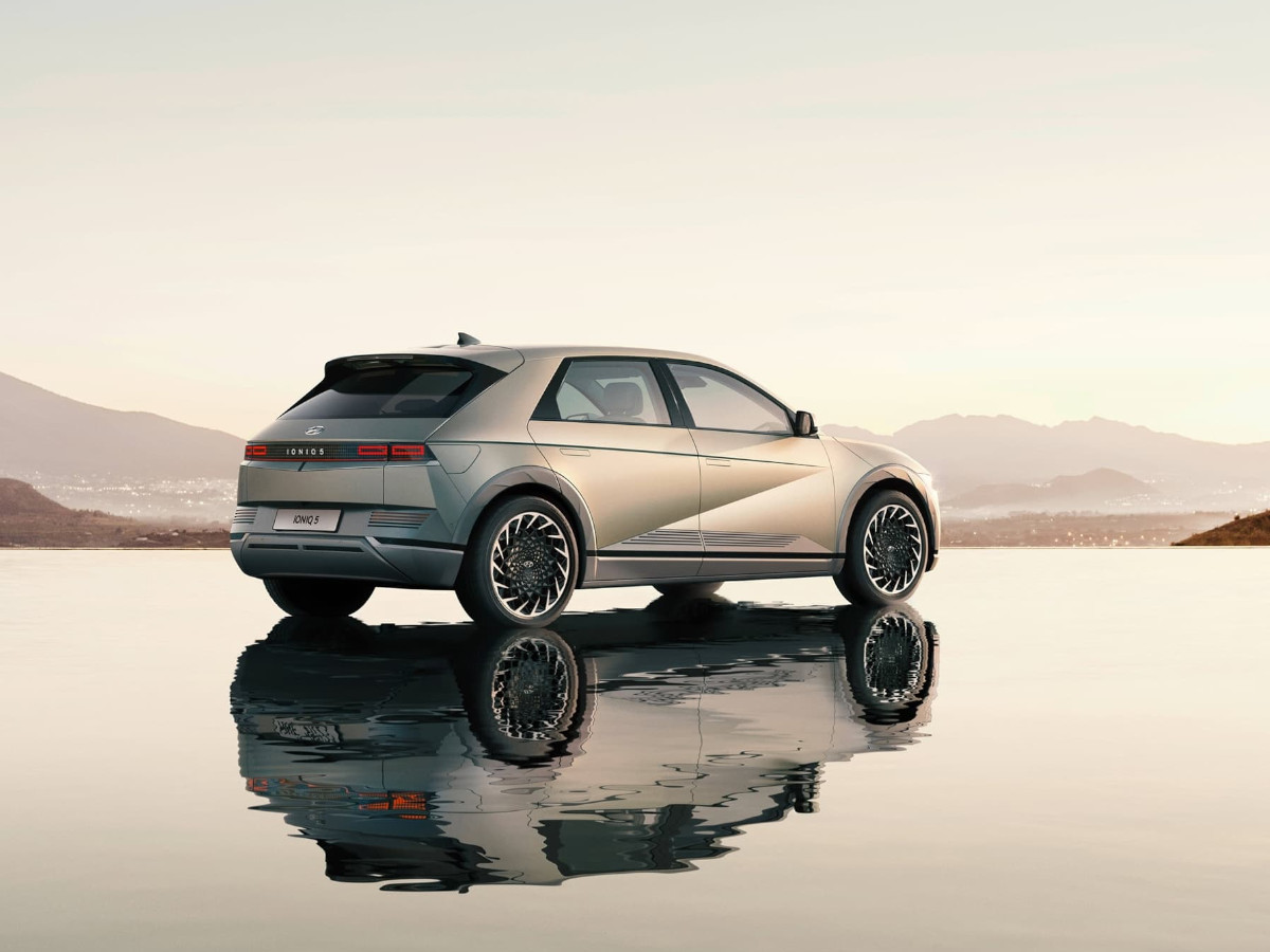 Hyundai Ioniq 5 de color cobre visto en tres cuartos traseros, estacionado sobre una superficie reflectante con un paisaje de montañas y cielo despejado al fondo.