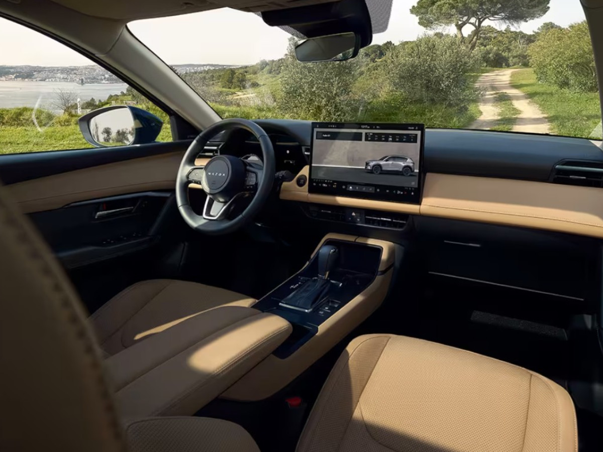 Interior del Mazda CX-5 con salpicadero en tonos beige y negros, volante Mazda, pantalla central y vista a un camino rural a través del parabrisas.