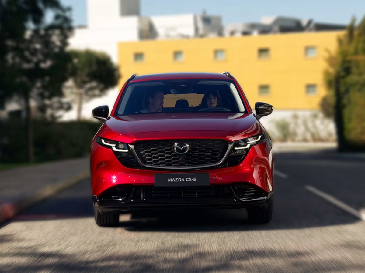Mazda CX-5 de color rojo visto de frente circulando por una calle urbana, con dos ocupantes visibles en el interior y edificios desenfocados al fondo.