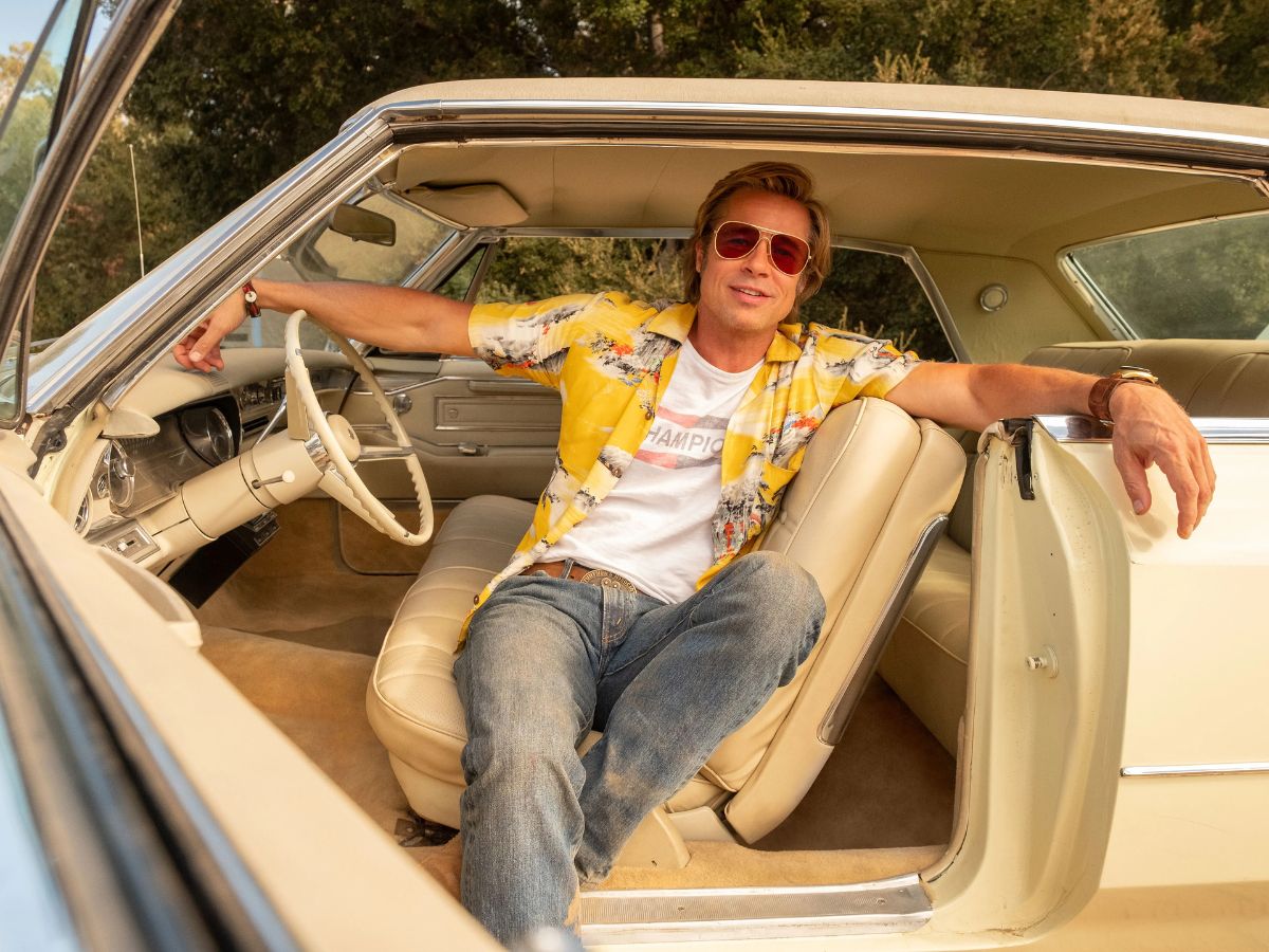 Brad Pitt con gafas de sol, una camisa de manga corta amarilla y unos vaqueros sentado dentro de un coche en una escena de la película Érase una vez en...Hollywood.