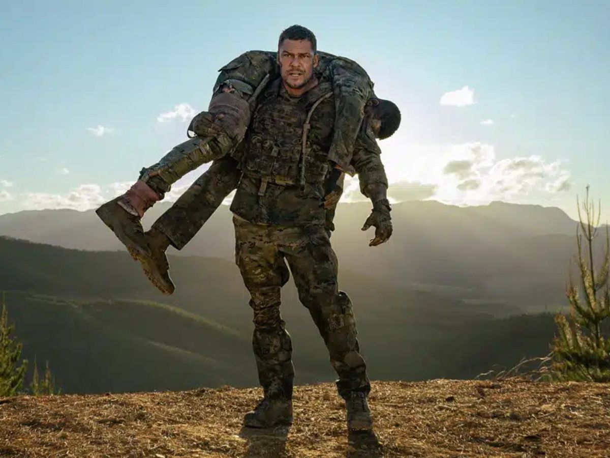 Alan Ritchson en el desierto vestido de militar cogiendo a alguien a hombros en la película Máquina de hierro.