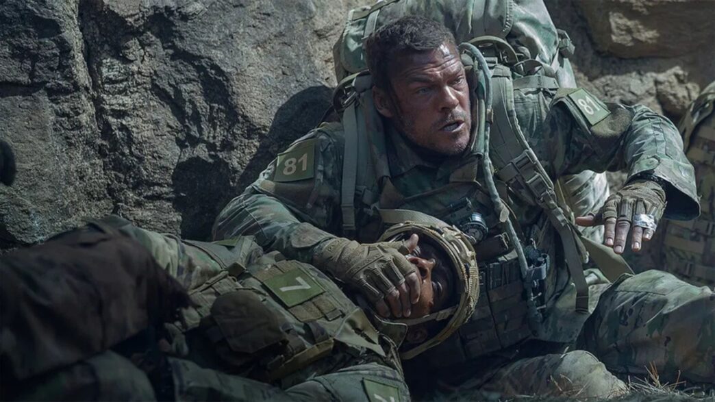 Alan Ritchson vestido de militar y tapando a otro soldado con la mano en una escena de la película Máquina de Hierro que triunfa en Netflix.