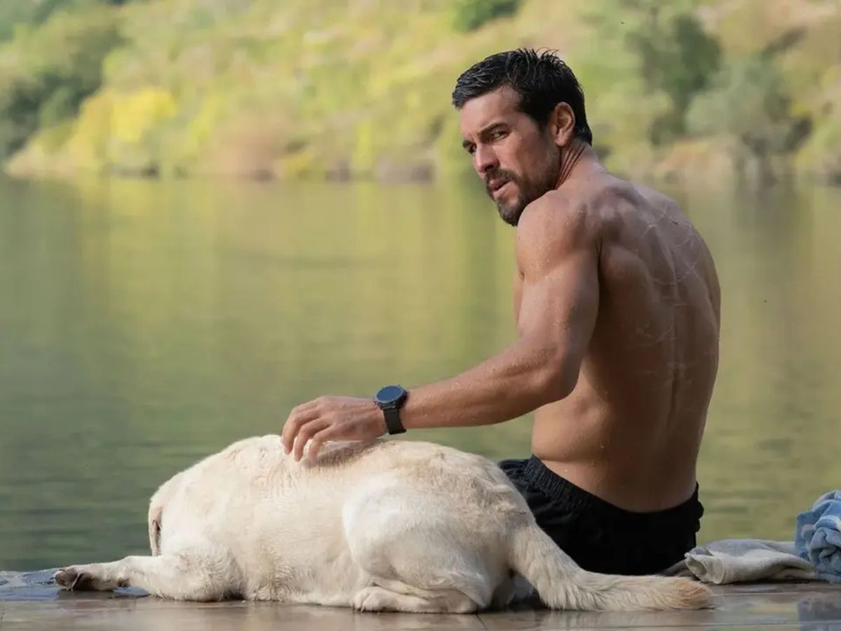 Mario Casas sentado en un muelle sin camiseta y acariciando a un perro blanco en la película Zeta.
