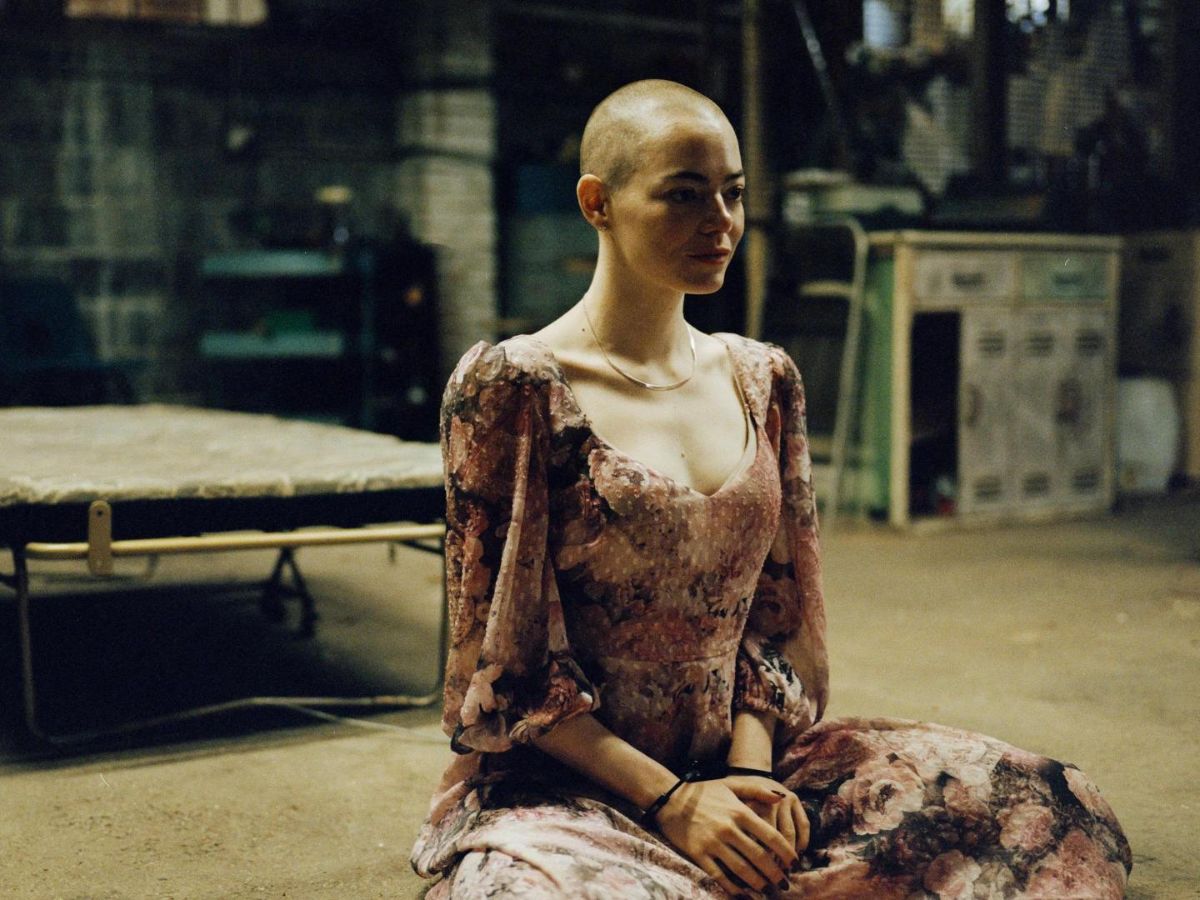 Emma Stone sin pelo sentada en el suelo con un vestido largo en una escena de la película Bugonia.