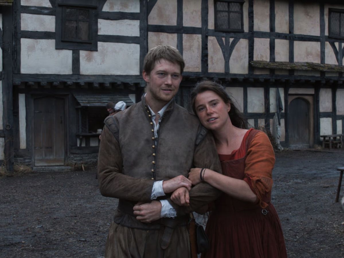 La actriz Jessie Buckley con un vestido rojo de época agarrada al brazo de Joe Alwyn, su hermano en la película Hamnet.