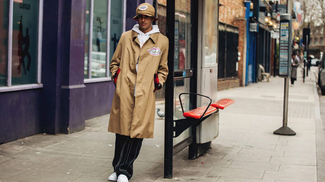 Hombre vestido con una gabardina beige y un sombrero apoyado en una parada de autobús en la calle