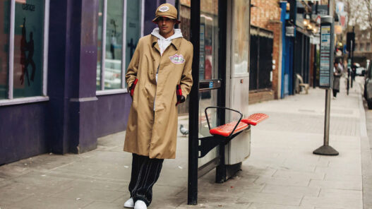 Hombre vestido con una gabardina beige y un sombrero apoyado en una parada de autobús en la calle