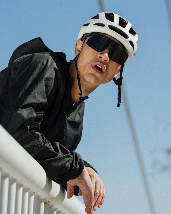 Hombre con casco blanco de ciclismo y gafas Mó Sport Fiber apoyado en una barandilla con el cielo azul de fondo