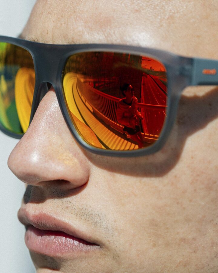 Primer plano de unas gafas Mó Sport Fiber con lente espejada roja que refleja a un corredor cruzando un puente