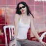 Milena Smit con top blanco y gafas de sol, sentada en sillas blancas en el stand Hennessy Clúb de los conciertos de Bad Bunny en Madrid
