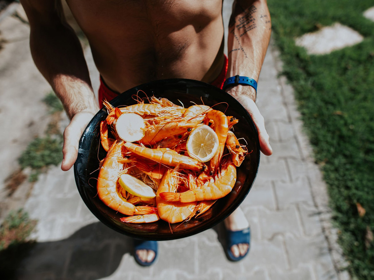 Hombre en bañador sostiene al sol un plato de langostinos con rodajas de limón en un jardín, en una escena de comida veraniega.