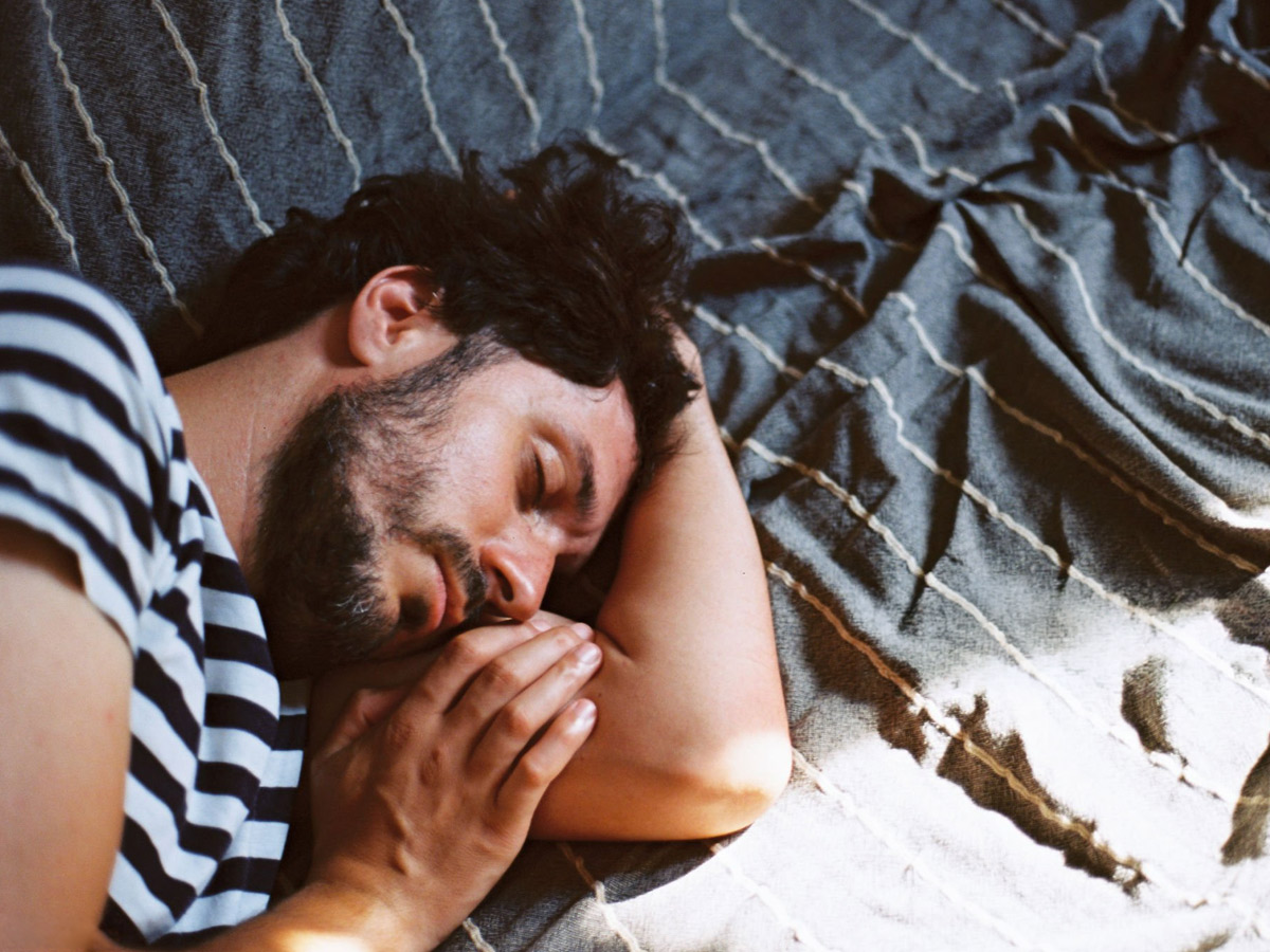 Hombre durmiendo de día sobre una colcha de rayas, con la luz cálida del verano entrando por la ventana.