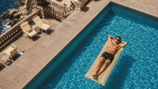 Hombre tumbado sobre una colchoneta en una piscina junto al mar, tomando el sol sin protección solar.