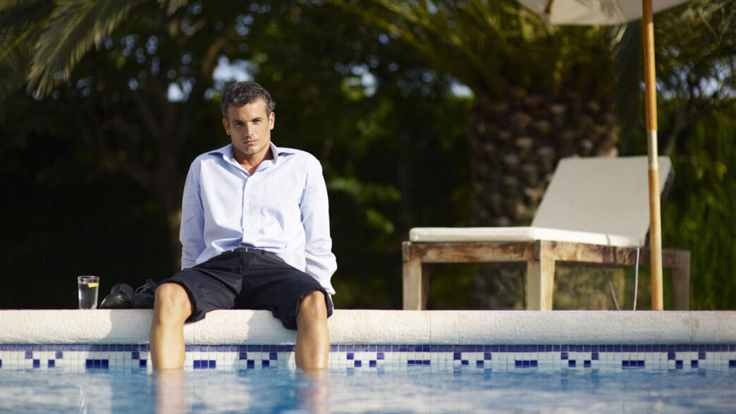 Hombre vestido con camisa y pantalón corto sentado al borde de una piscina con los pies en el agua y gesto pensativo