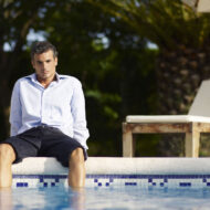 Ni es el calor ni el aburrimiento: por qué el verano dispara el estrés en las personas más organizadas, según una psiquiatra Hombre vestido con camisa y pantalón corto sentado al borde de una piscina con los pies en el agua y gesto pensativo
