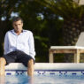 Hombre vestido con camisa y pantalón corto sentado al borde de una piscina con los pies en el agua y gesto pensativo