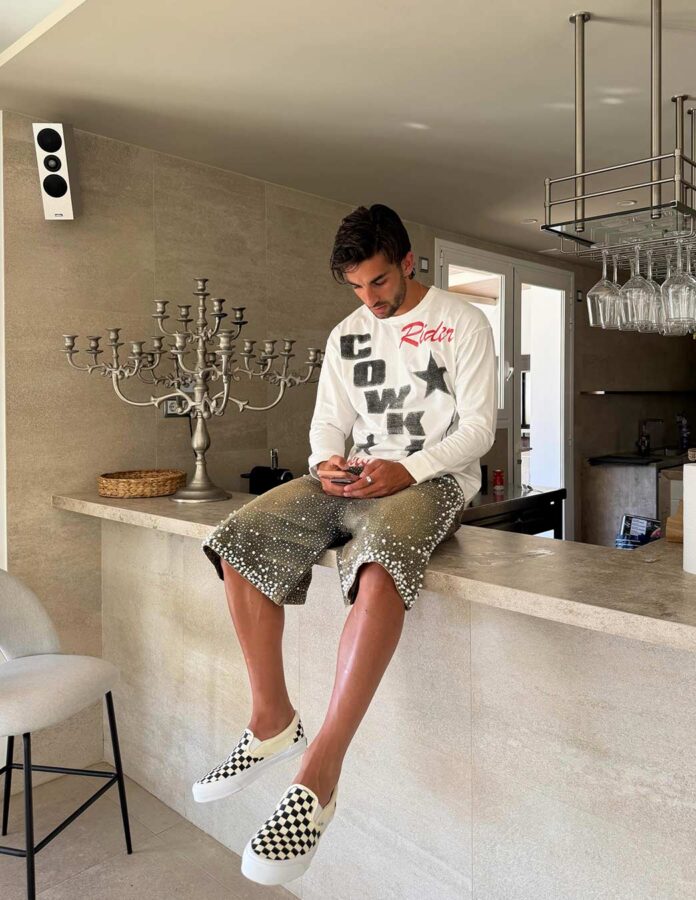Ferran Torres sentado en una barra de cocina con bermudas vaqueras bordadas con perlas y zapatillas de cuadros