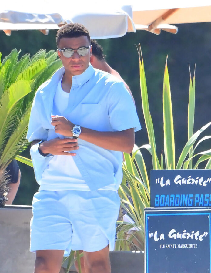 Kylian Mbappé con conjunto azul celeste y gafas de espejo junto a los carteles del restaurante La Guérite en la Île Sainte-Marguerite