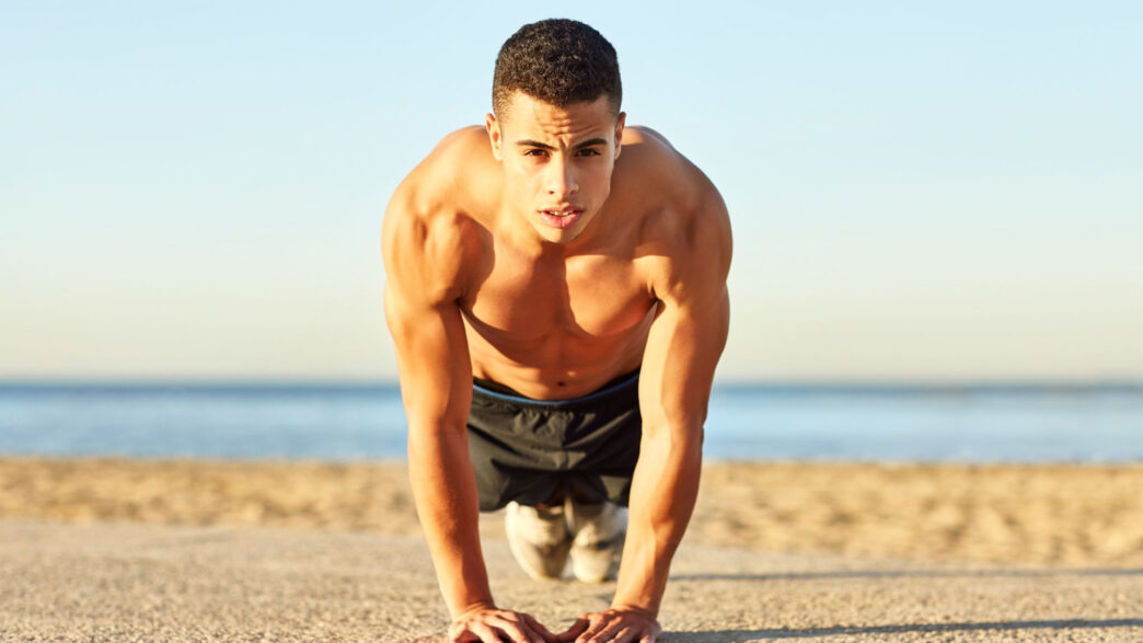 Hombre haciendo flexiones en la playa para entrenar el pectoral en verano sin material