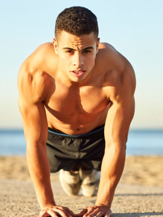 Hombre haciendo flexiones en la playa para entrenar el pectoral en verano sin material