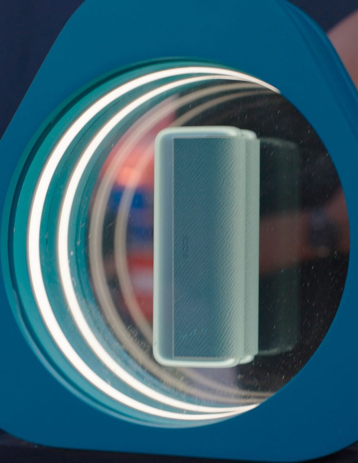 Dispositivo IQOS expuesto en una vitrina circular con anillos de luz blanca en la House of IQOS