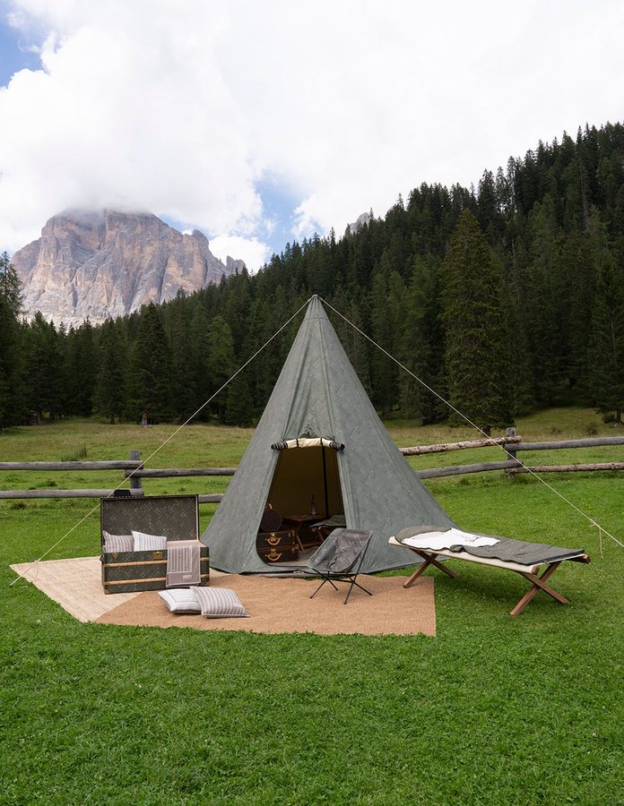 Tienda de campaña, baúl y camastro Louis Vuitton en un prado de los Dolomitas con la montaña al fondo