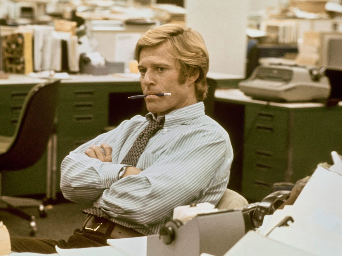 Robert Redford, vestido con una camisa a rayas azules y blancas y una corbata estampada, con los brazos cruzados y un lápiz en la boca, en una recreación de las oficinas de «The Washington Post» en la película «Todos los hombres del presidente»