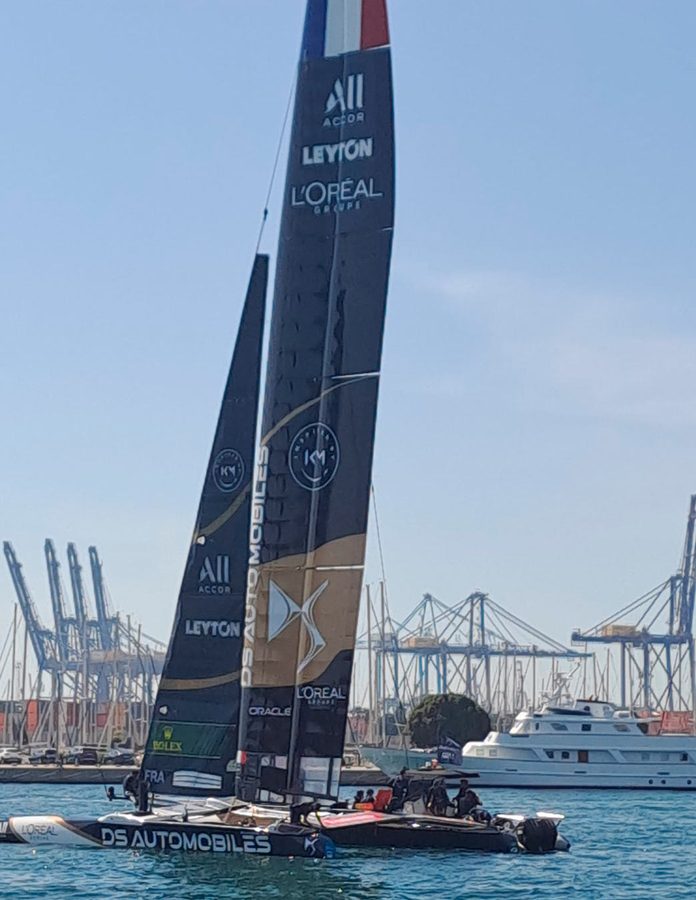 Catamarán del DS Automobiles SailGP Team France junto a una embarcación auxiliar, con grúas portuarias al fondo.