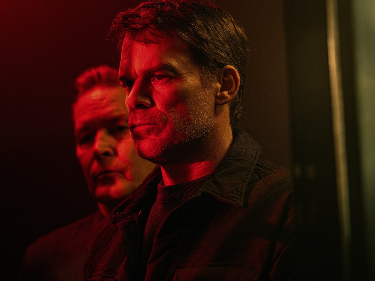 Michael C. Hall, como Dexter Morgan, de perfil bajo una luz roja con un hombre mayor detrás en la segunda temporada de Dexter: Resurrección