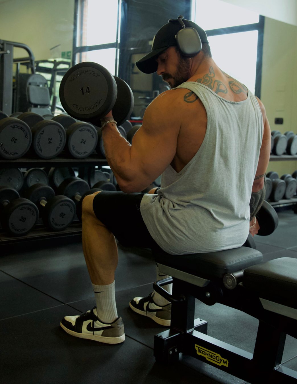 Hombre sentado en un banco de gimnasio haciendo curl con una mancuerna de 14 kilos frente a un soporte de mancuernas Technogym