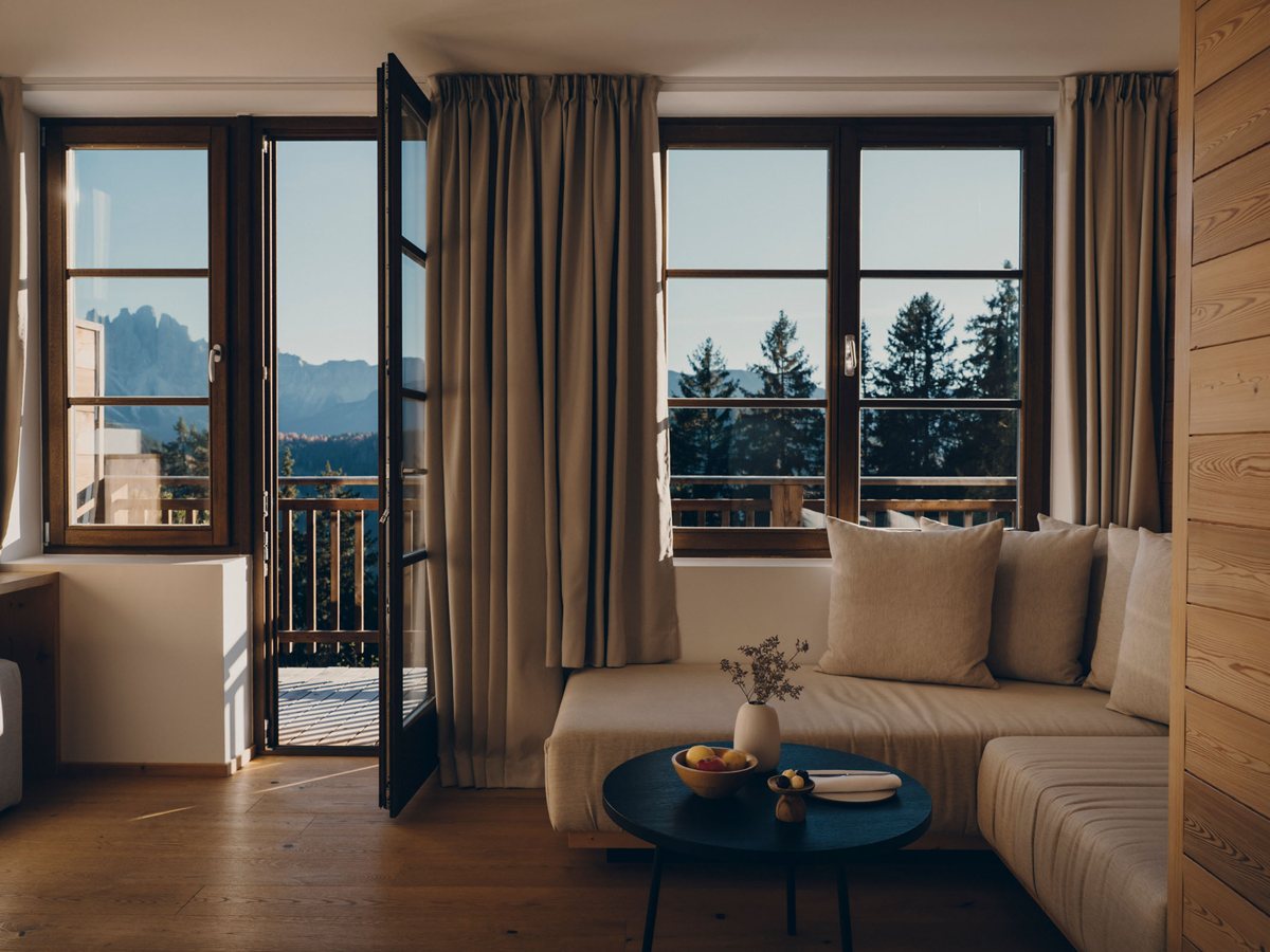 Salón de una suite del edificio histórico de FORESTIS con sofá, puerta abierta a la terraza de madera y los Dolomitas al fondo