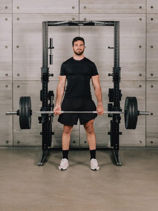 Hombre de pie con la espalda recta sujetando una barra cargada delante de un rack Technogym