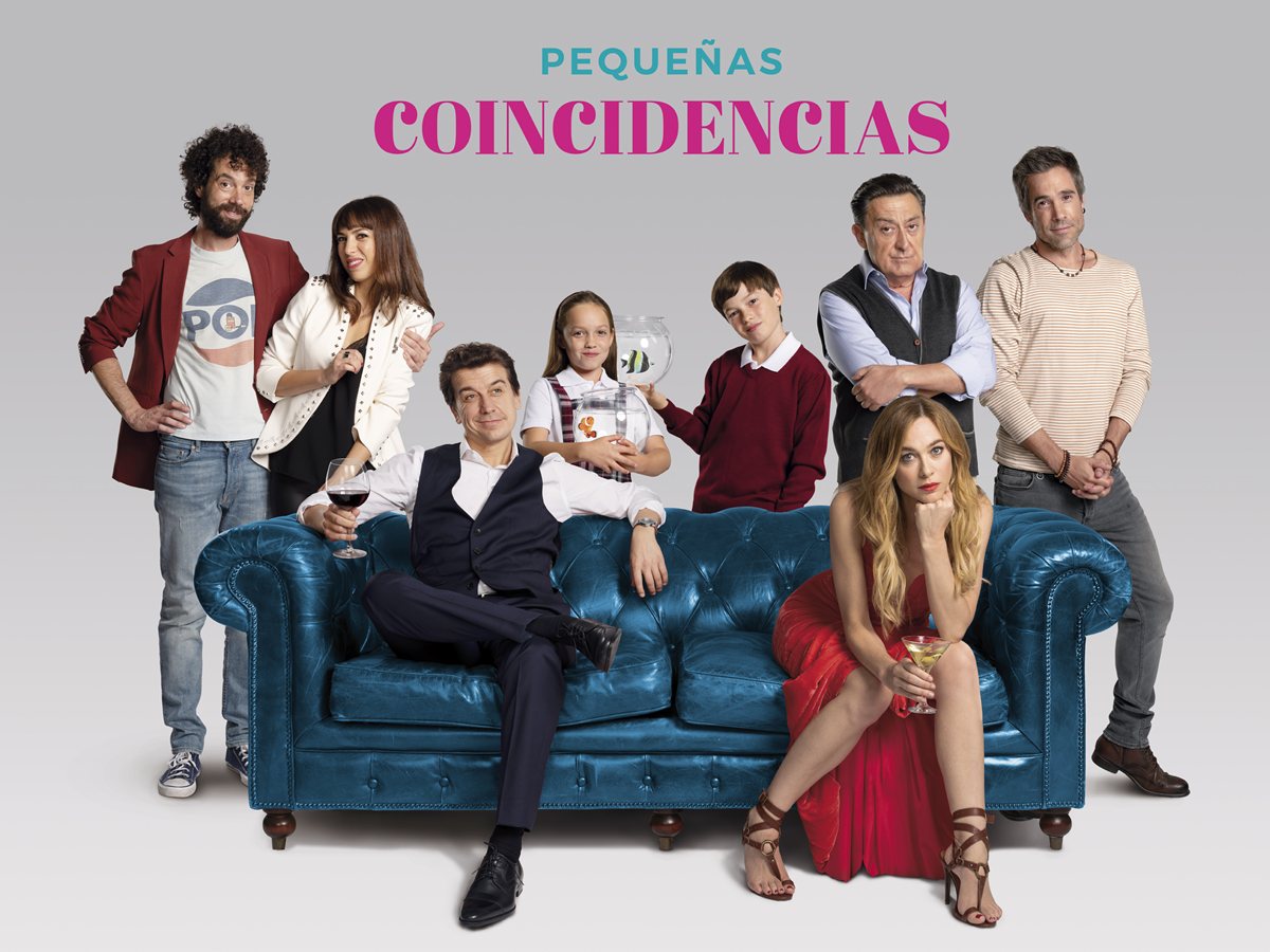 Javier Veiga, con una copa de vino, y Marta Hazas, de rojo, sentados en un sofá azul con el resto del reparto de Pequeñas coincidencias y una niña con una pecera