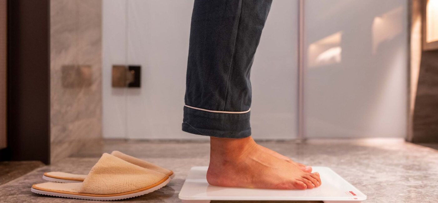 Hombre en pijama subido a una báscula de baño junto a unas zapatillas de casa, con la cintura fuera de plano