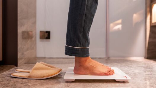 Hombre en pijama subido a una báscula de baño junto a unas zapatillas de casa, con la cintura fuera de plano