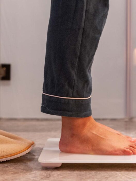 Hombre en pijama subido a una báscula de baño junto a unas zapatillas de casa, con la cintura fuera de plano