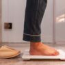 Hombre en pijama subido a una báscula de baño junto a unas zapatillas de casa, con la cintura fuera de plano