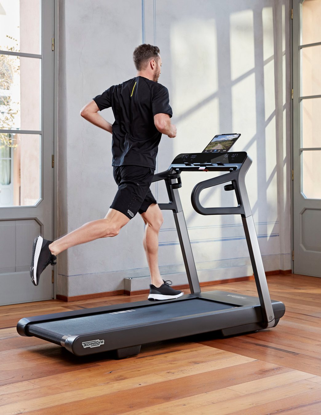 Hombre corriendo en una cinta Technogym en un salón con suelo de madera y ventanales