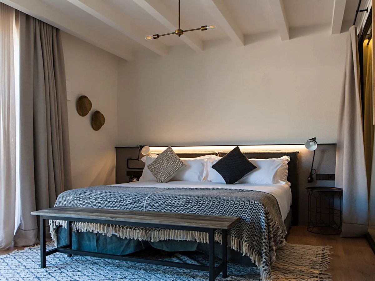 Dormitorio de Terra Dominicata con cama de 200 por 200, vigas blancas, cabecero retroiluminado y banco de madera a los pies