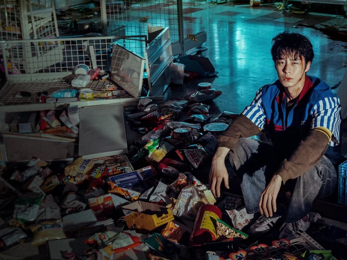 Lee Jae-wook, en cuclillas y con uniforme de tienda, entre estanterías volcadas y paquetes por el suelo en Un trabajo maldito
